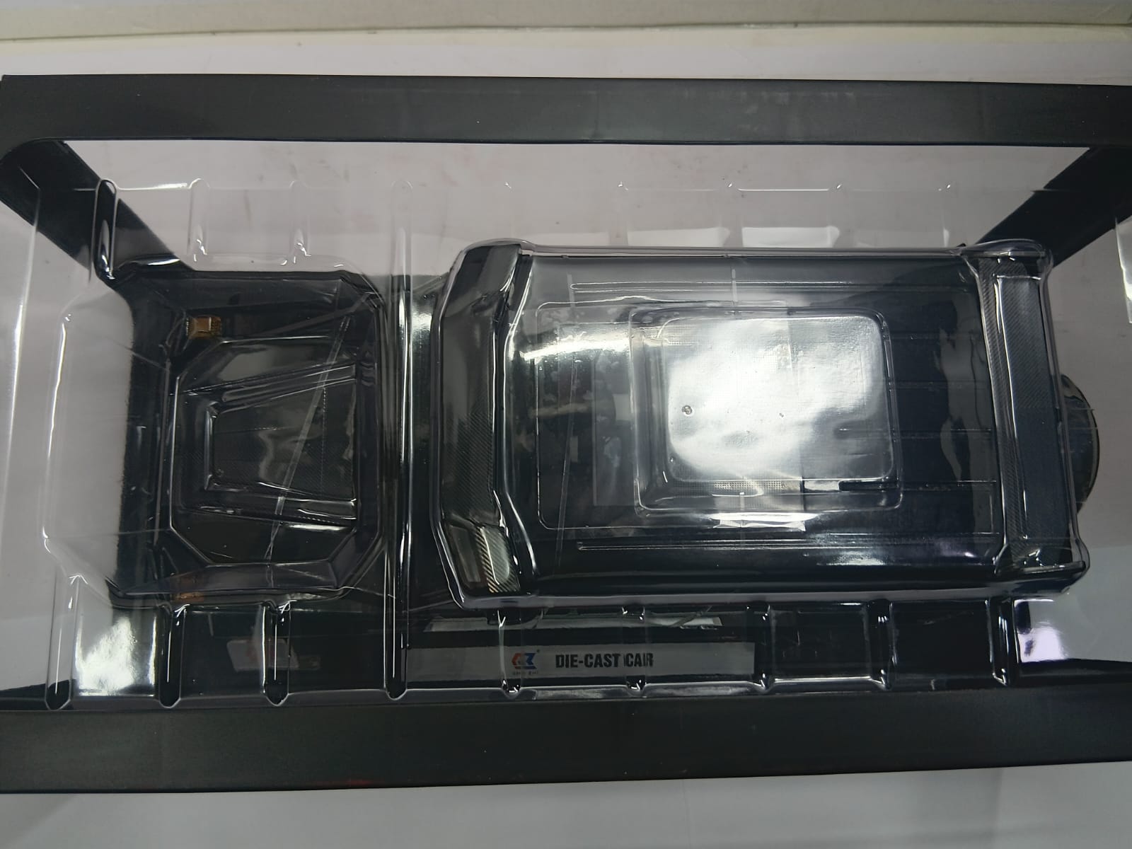 A black die-cast car part, likely a bumper or fender section, packaged in a clear plastic protective sleeve inside a black tray.