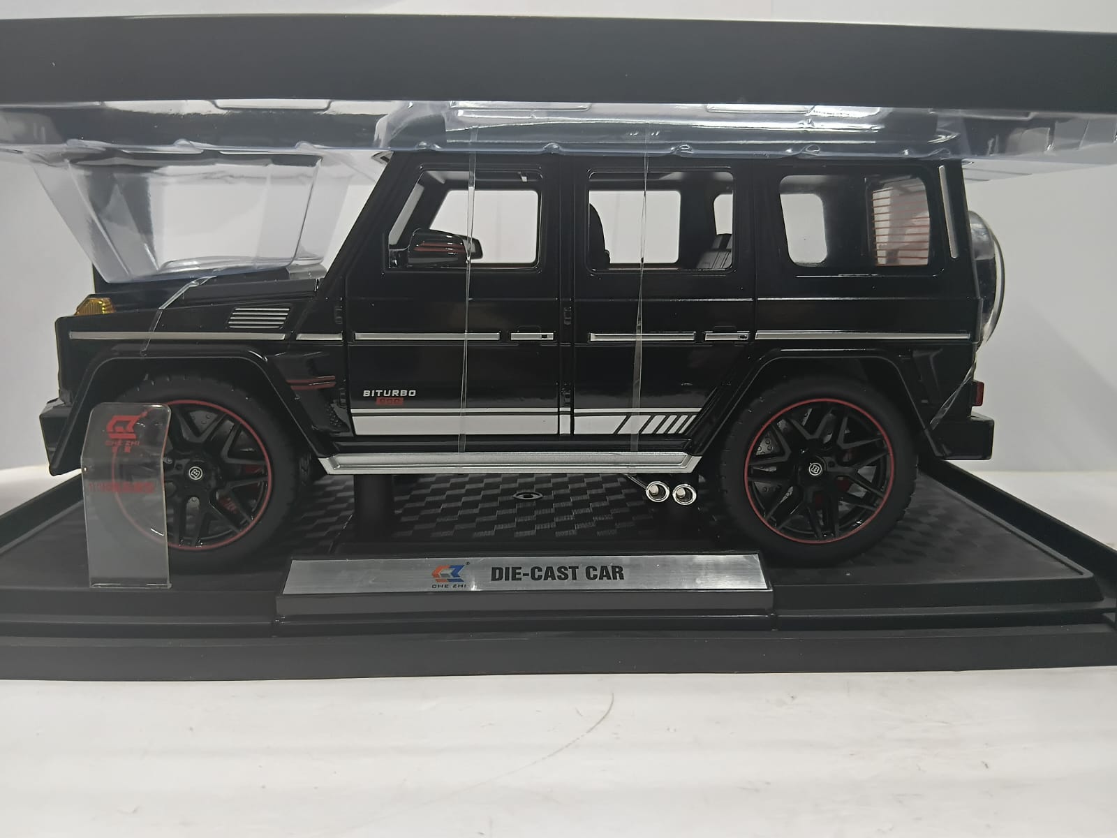 A black die-cast model car of a Mercedes-Benz G-Class with silver stripes and red wheel accents, displayed on a black base inside a clear plastic case.